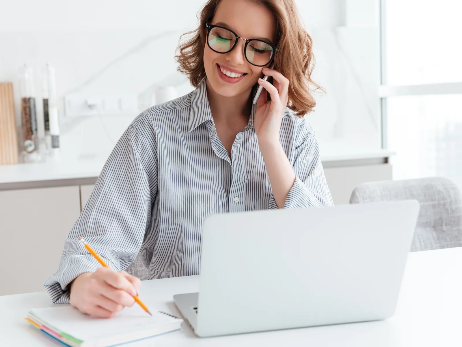 Consultora de óculos ao telefone, anotando num caderno ao lado do notebook